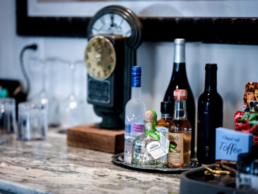 Um balcão de bar com diversas garrafas de bebidas alcoólicas, copos e um relógio.