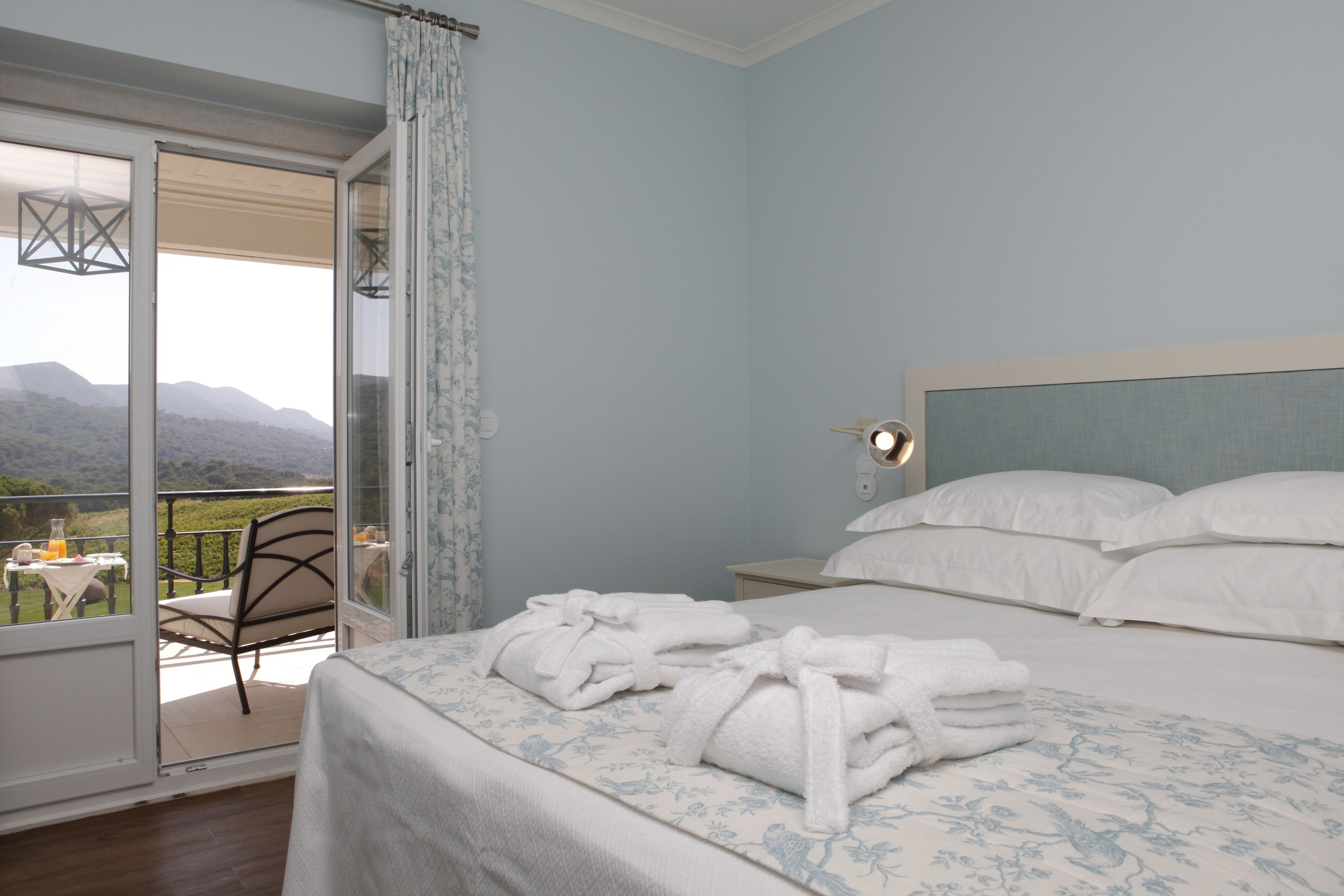 A bedroom with a bed, white pillows, and a nightstand. There is a window with a view of mountains and a table with a chair outside.