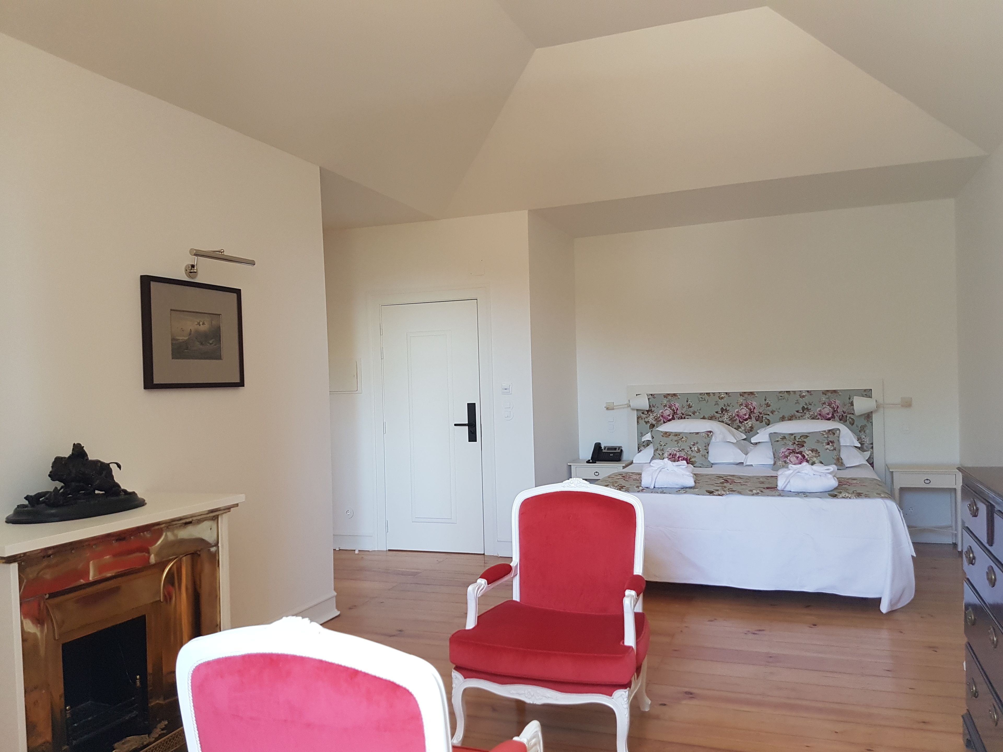 A hotel room with a wooden floor, a bed with floral bedding, a red chair, a white door, and a fireplace.
