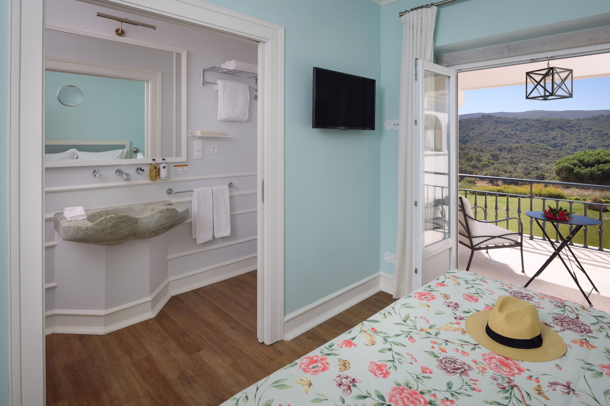 A bedroom with a stone sink, a flat-screen TV, and a balcony with a view of the hills.