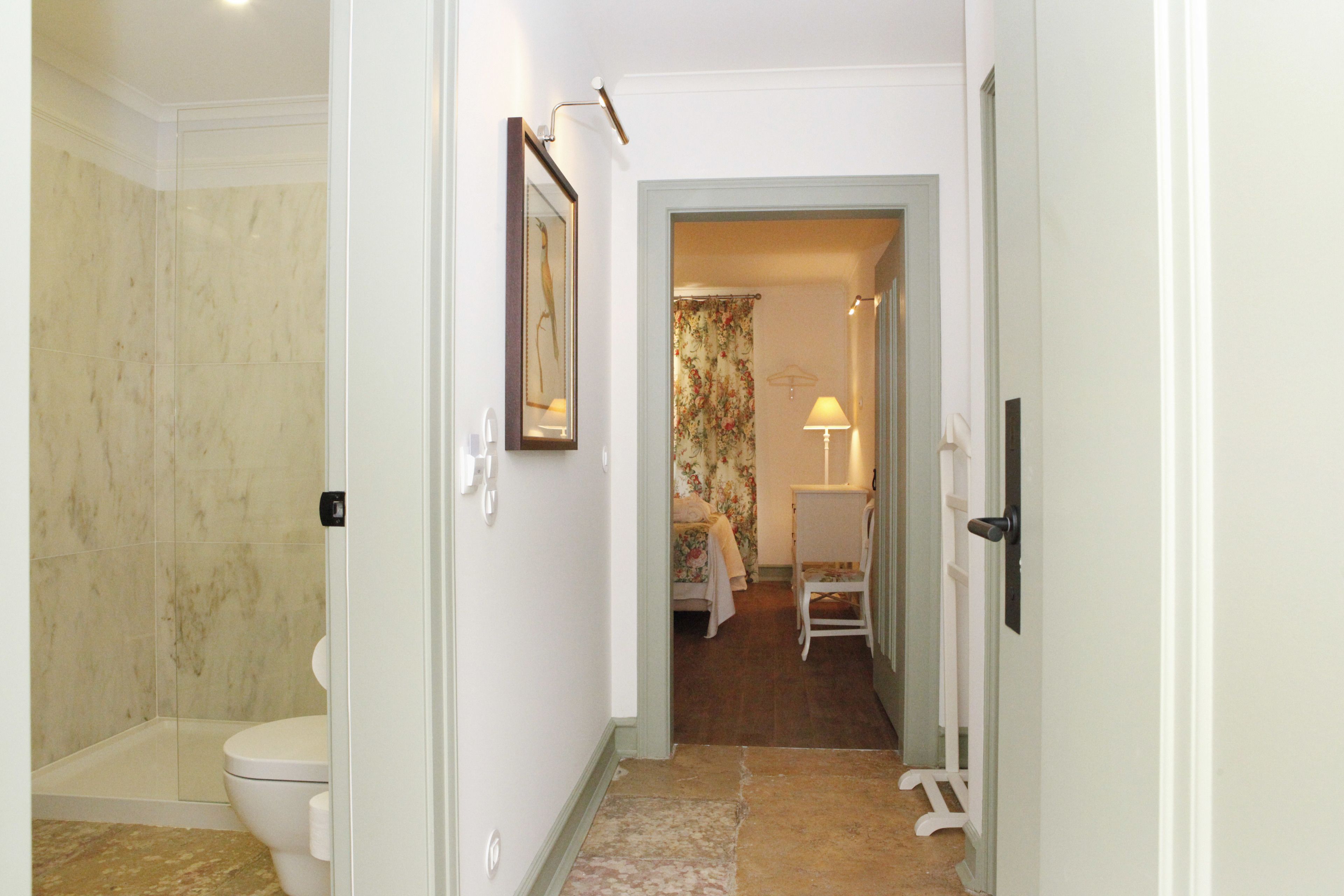 A bathroom with a toilet, shower, and a hallway leading to a bedroom with a bed and a nightstand.