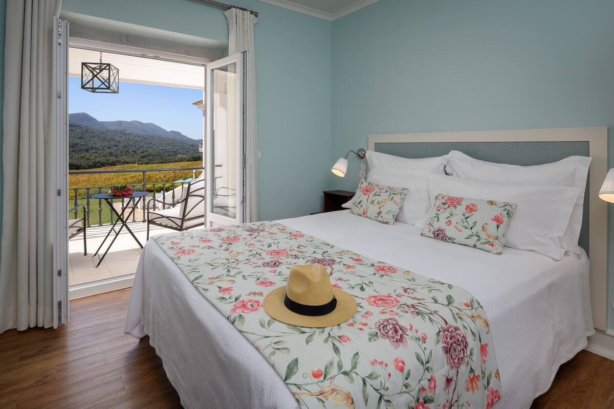 A bedroom with a bed, floral bedding, a hat on the bed, a nightstand, a lamp, and a view of a balcony with a table and chairs.