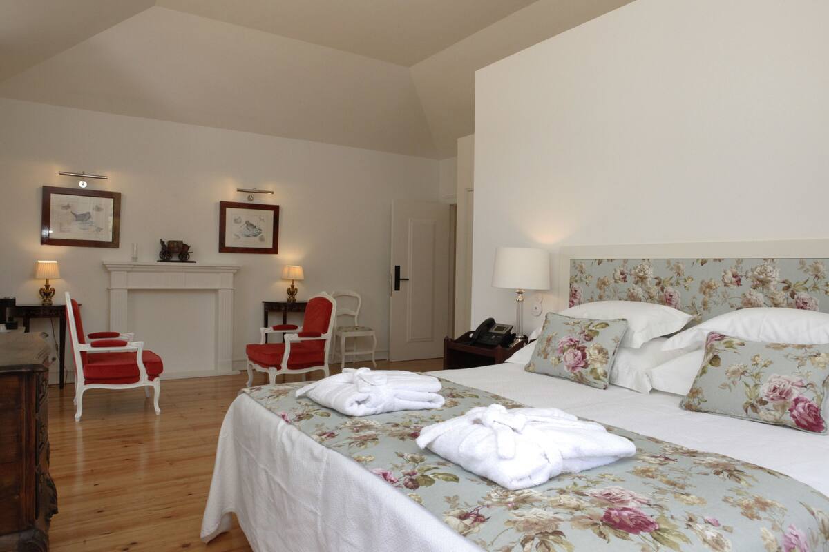 A hotel room with a bed, two chairs, a fireplace, and framed pictures on the wall.