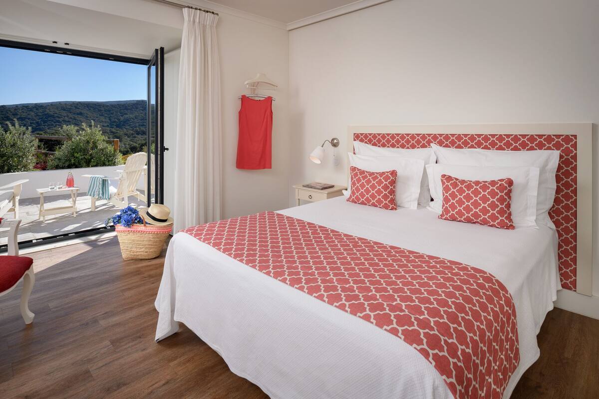 A bedroom with a large bed, a nightstand, a lamp, a chair, a table, and a view of the outdoors.