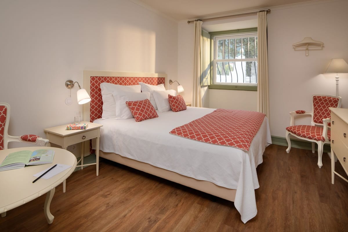 A neatly arranged bedroom with a large bed, bedside tables, a chair, and a small table with books and a pen.