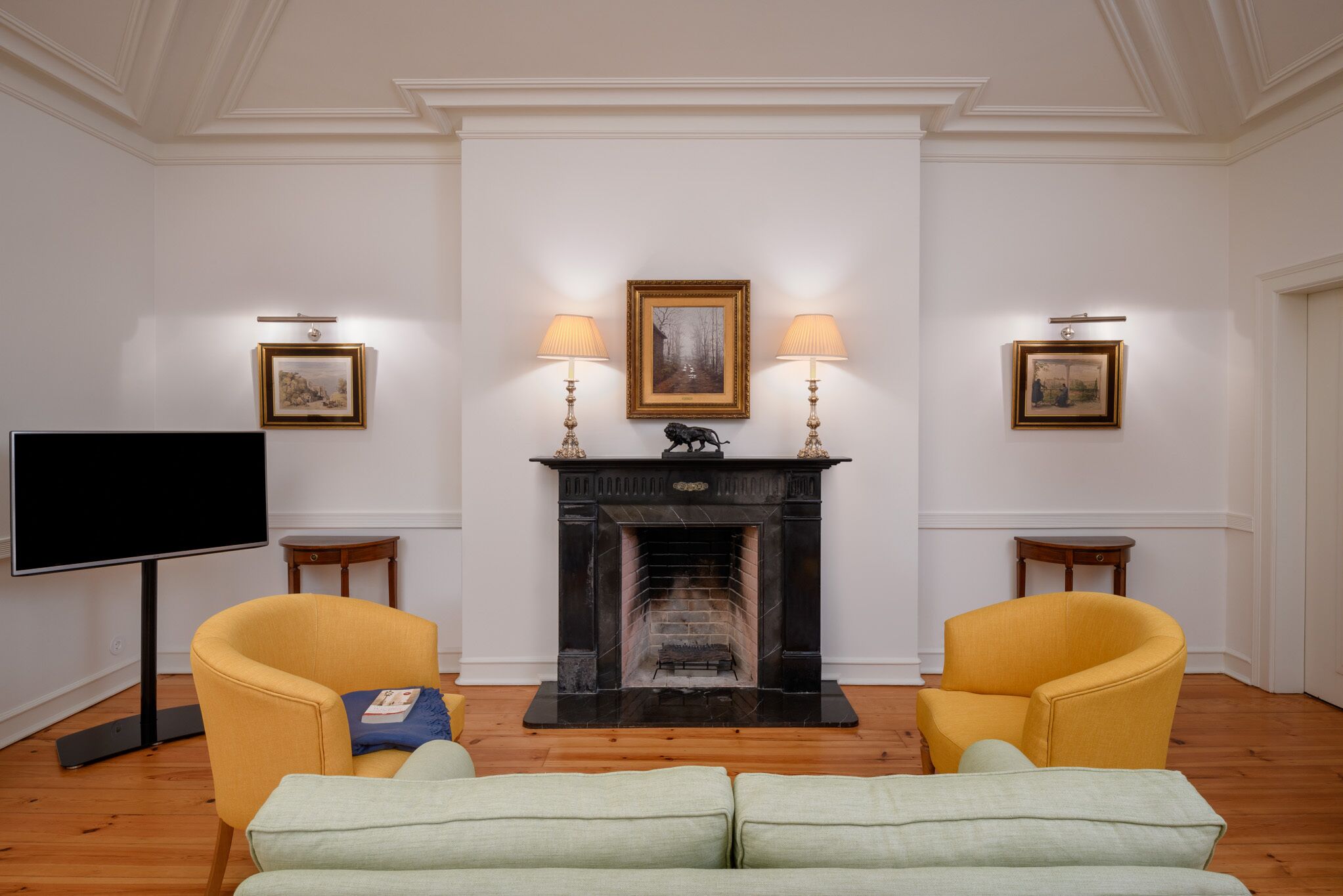 Una sala de estar con chimenea, dos sillones amarillos, un sofá verde, un televisor de pantalla plana y cuadros enmarcados en las paredes.