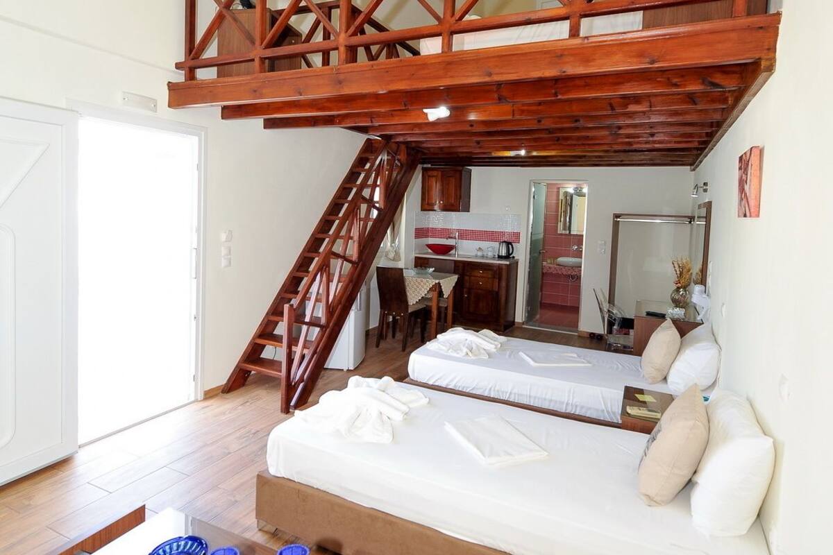 A loft-style bedroom with a wooden truss ceiling, a staircase, a bed, a desk, and a bathroom in the background.