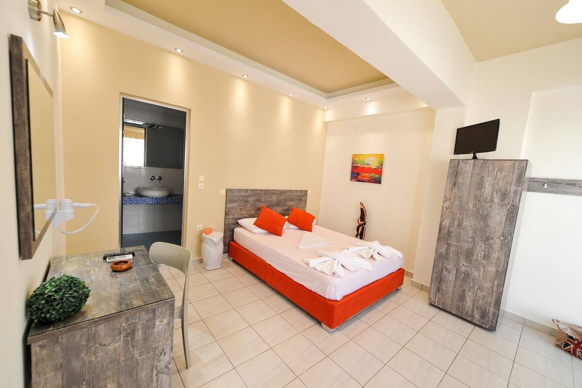 A hotel room with a bed, a desk, a chair, a TV, and a bathroom visible through an open door.
