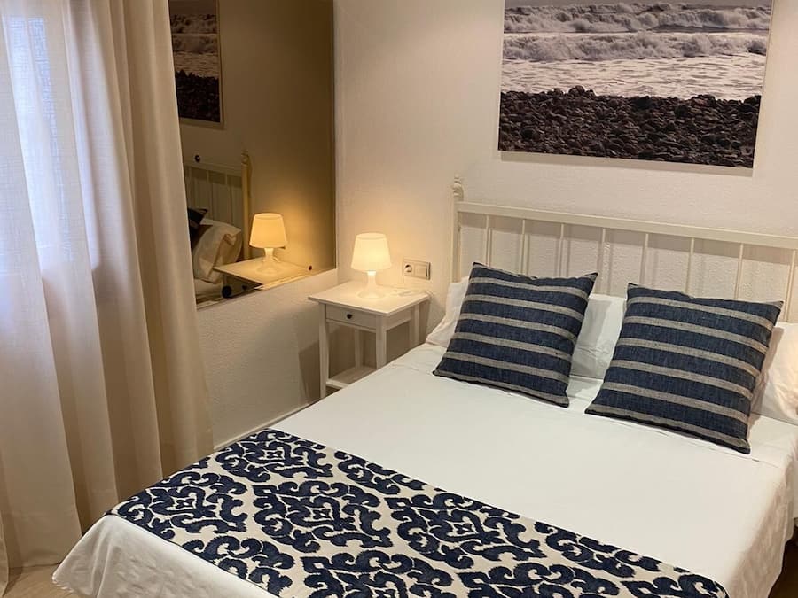 Una cama bien hecha con una colcha estampada, una mesita de noche con una lámpara, un cuadro en la pared que representa una costa rocosa y una ventana con cortinas.