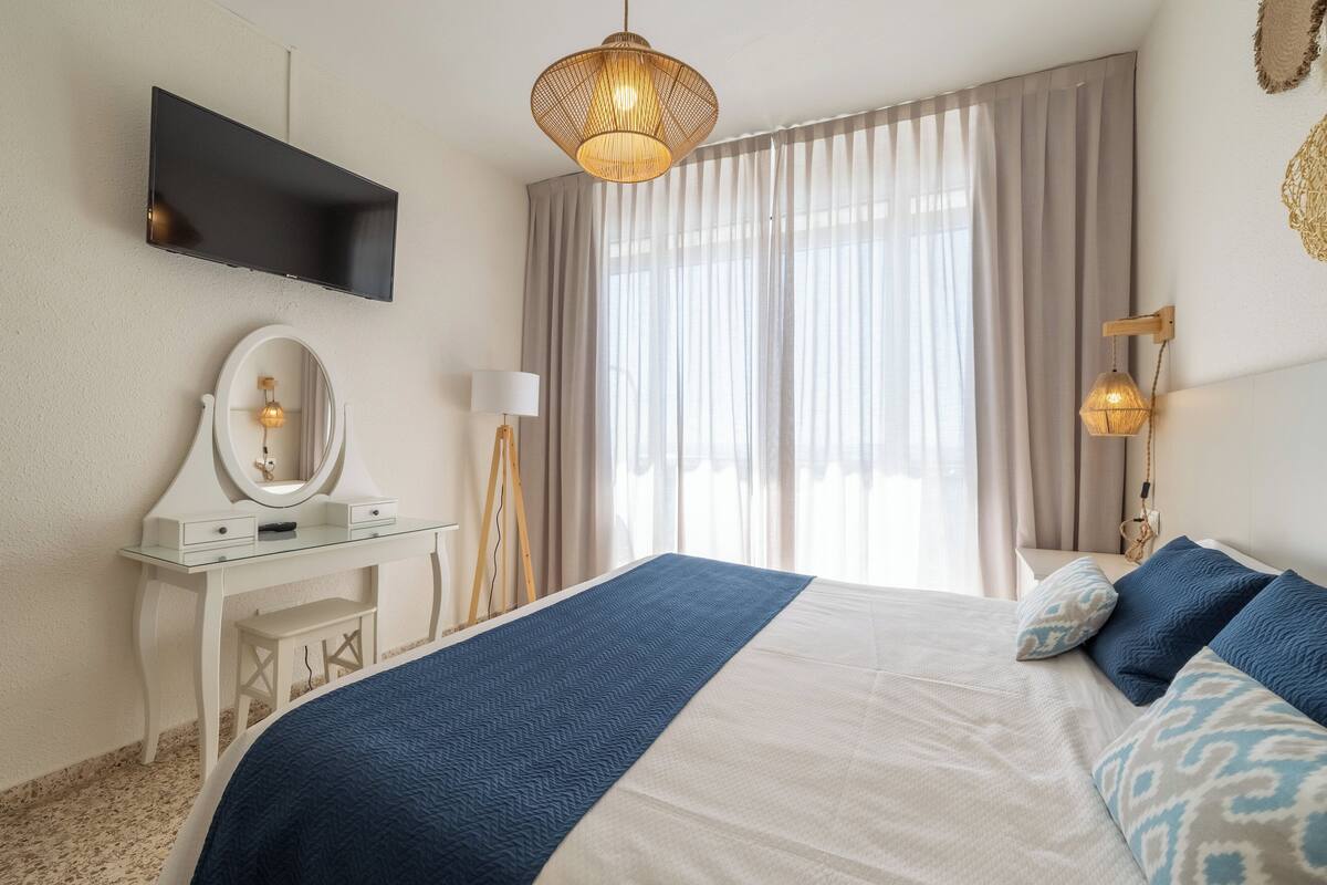 A bedroom with a bed, a vanity with a mirror, a flat-screen TV, and a large window with curtains.