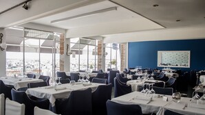 A restaurant with multiple tables, each set with cutlery and wine glasses, large windows offering a view of the marina, and a blue accent wall with a framed picture. - Hotel Ampolla Sol (L'Ampolla)