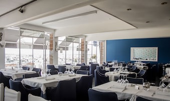 Un restaurante con varias mesas, cada una con cubiertos y copas de vino, amplias ventanas que dan a vista de la marina y una pared azul con un cuadro enmarcado.