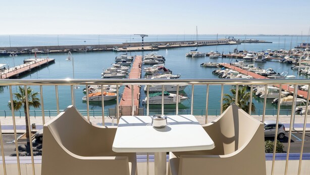 A balcony with two white chairs and a table, overlooking a marina with boats and a pier. - Hotel Ampolla Sol (L'Ampolla)