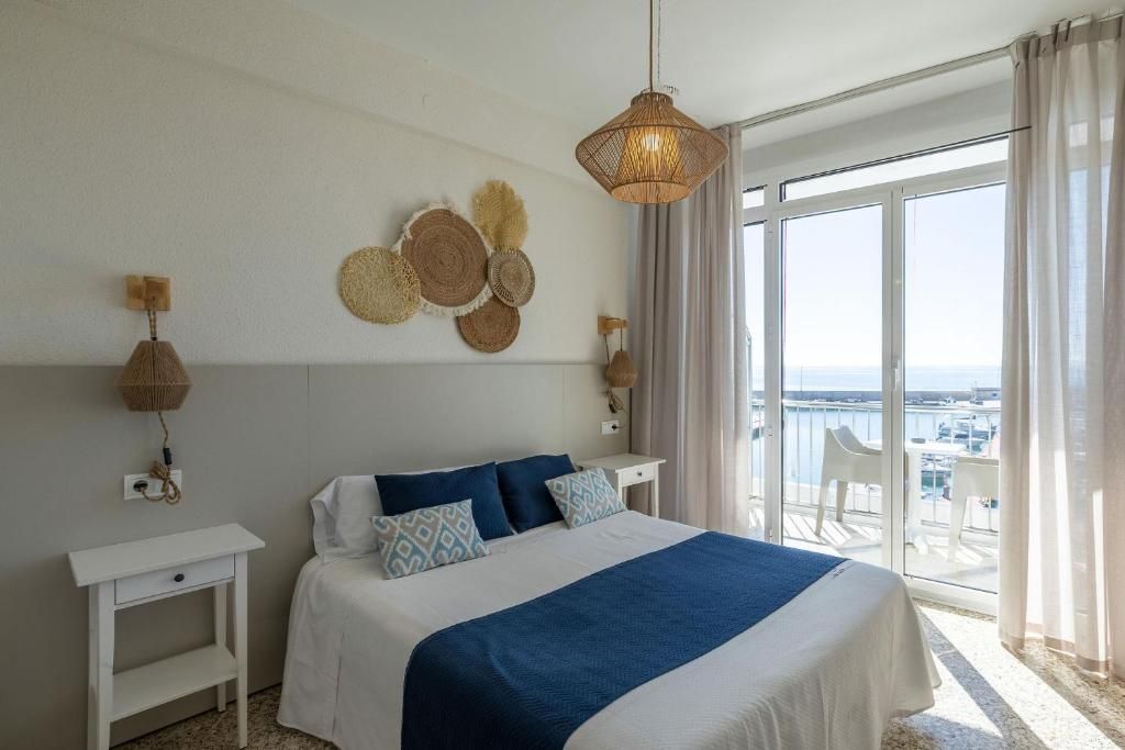A hotel room with a bed, a bedside table, a wall-mounted lamp, and a view of the sea through a large window.