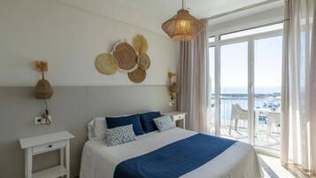 A hotel room with a bed, a bedside table, a wall-mounted lamp, and a view of the sea through a large window.