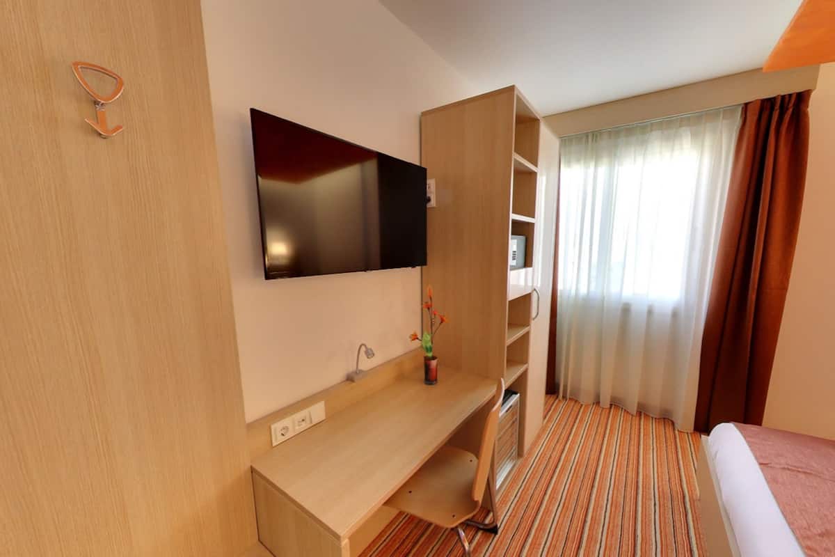 A hotel room with a bed, a desk, a TV mounted on the wall, a shelf unit, and a window with curtains.