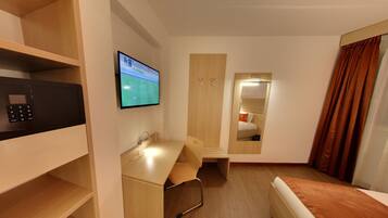 Habitación de hotel con escritorio, televisor montado en la pared, espejo, armario y cama con cortinas.