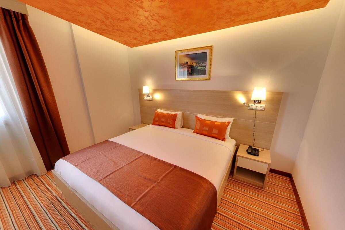 A hotel room with a large bed, two bedside lamps, a nightstand, and a cityscape picture on the wall.