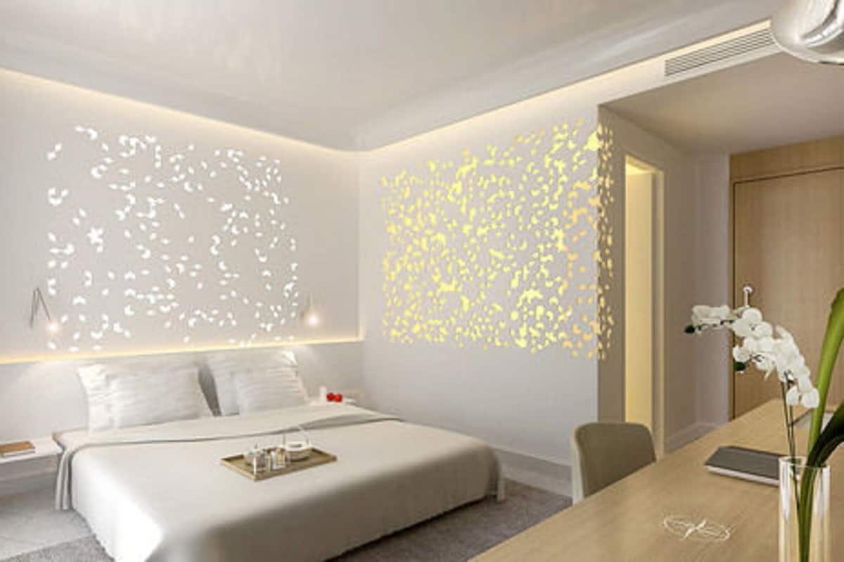 A modern hotel room with a large bed, a desk, and a chair. There is a decorative light installation on the wall and a vase with flowers on the desk.