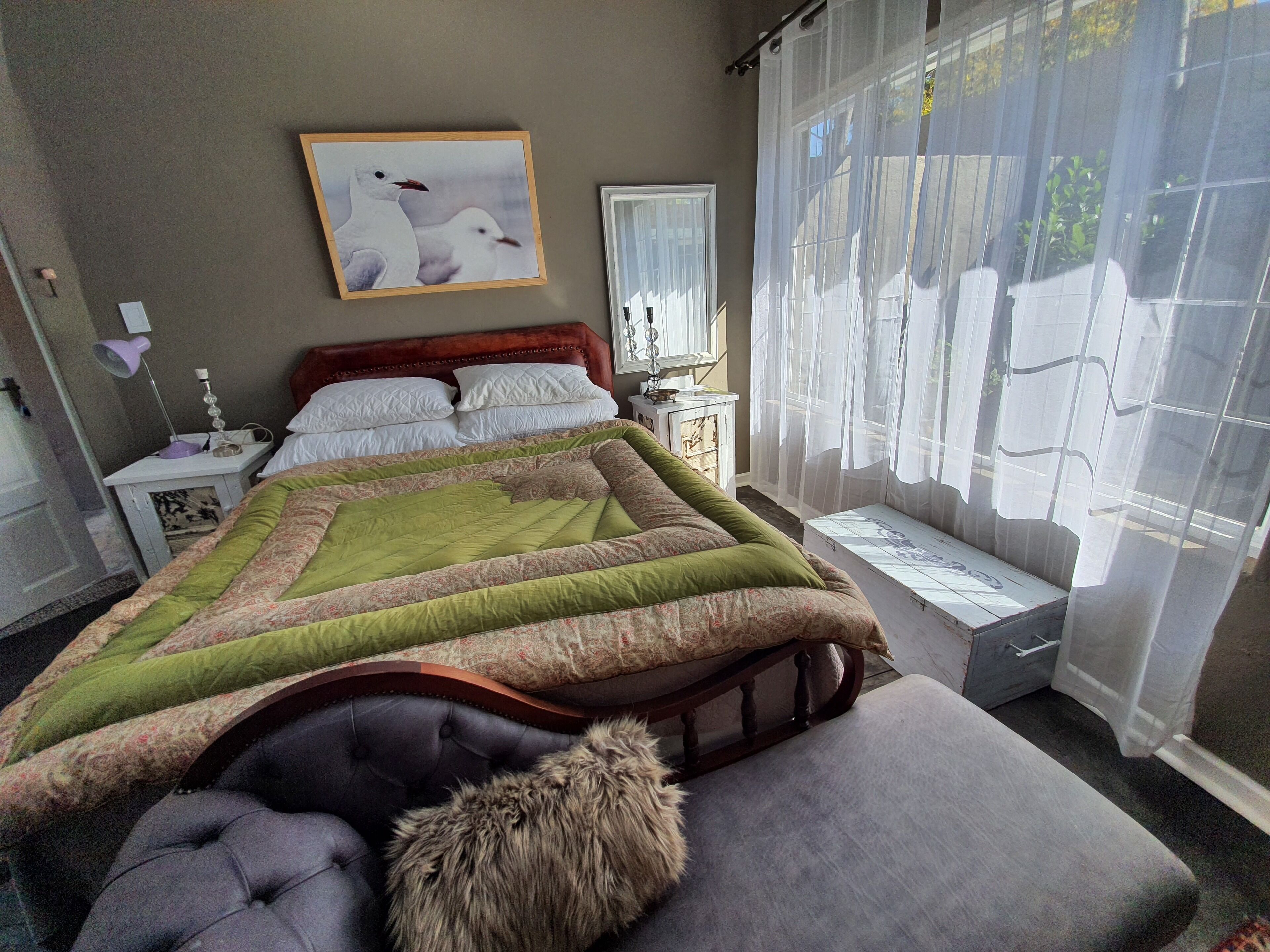 Une chambre avec un grand lit, une table de chevet avec une lampe, une fenêtre avec des rideaux légers et un tableau encadré au mur.