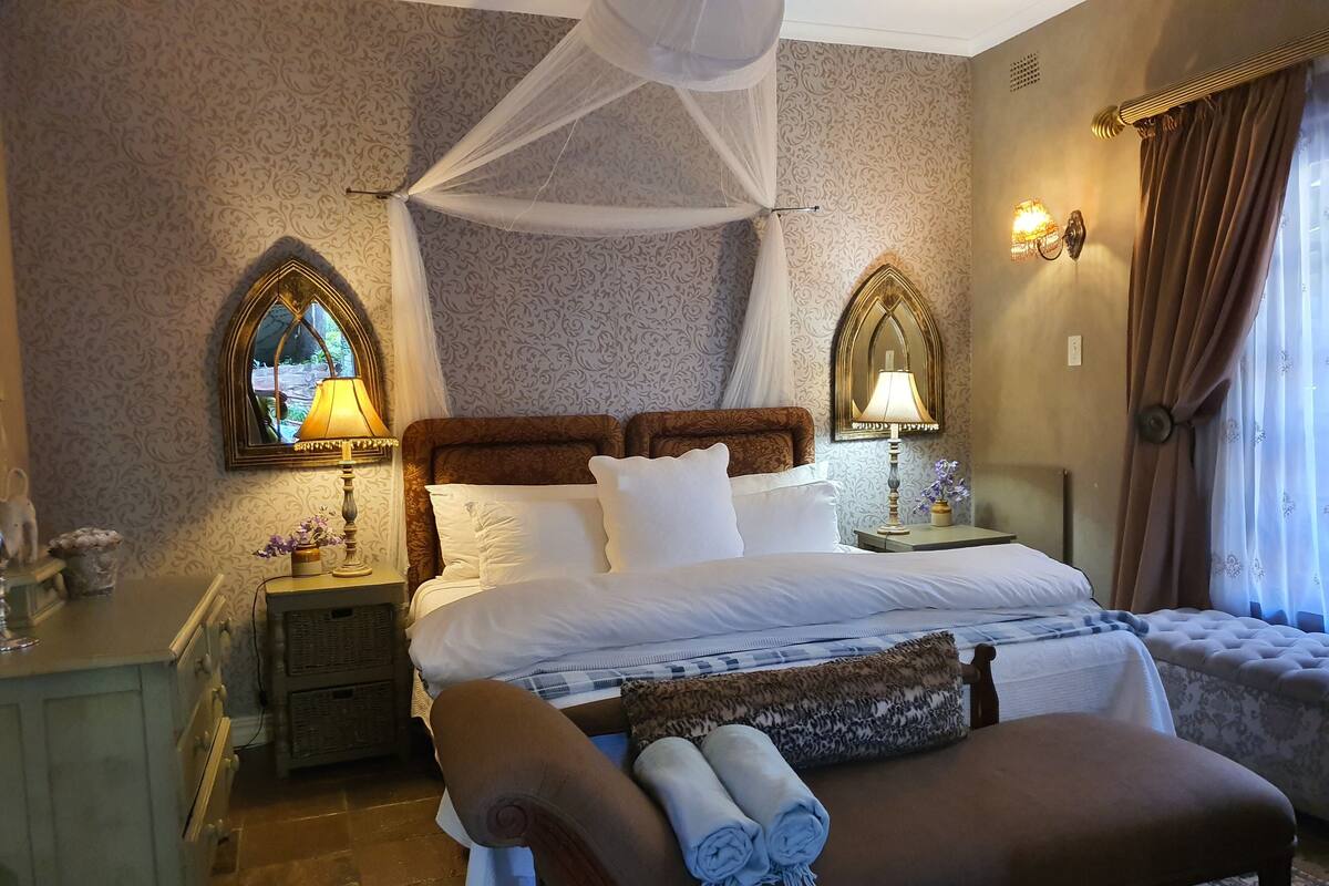 A bedroom with a canopy bed, two bedside lamps, a nightstand with a lamp, a bench with rolled towels, and a mirror on the wall.