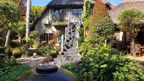 A house with a thatched roof, a staircase leading to a balcony, and a garden with a water feature. - Waterhouse Guest Lodges 202 Lawley Street (Pretoria)
