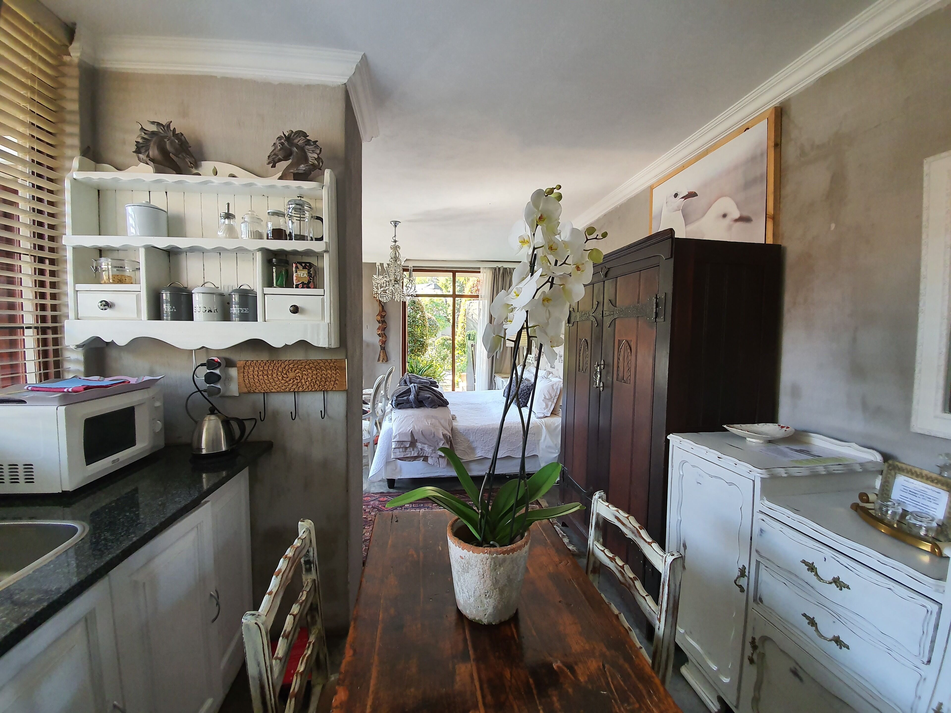 A narrow kitchen with a wooden table, a potted orchid, and a bed visible through an open door.