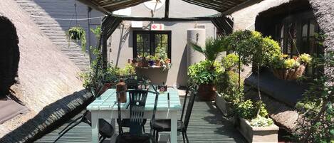 A covered outdoor seating area with a table and chairs, surrounded by potted plants and a thatched roof structure.