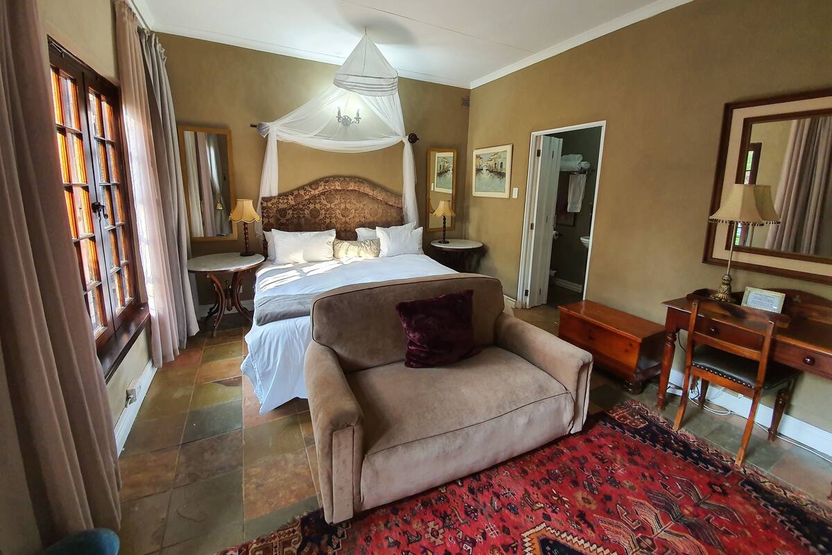 A bedroom with a large bed, a bedside table, a chair, a desk, and a mirror.
