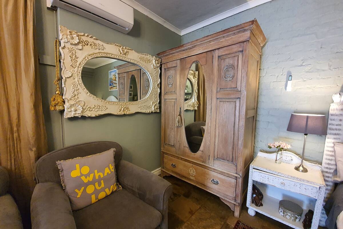 A room with a large ornate mirror, a brown armchair with a cushion, a wooden cabinet, and a small white side table with a lamp.