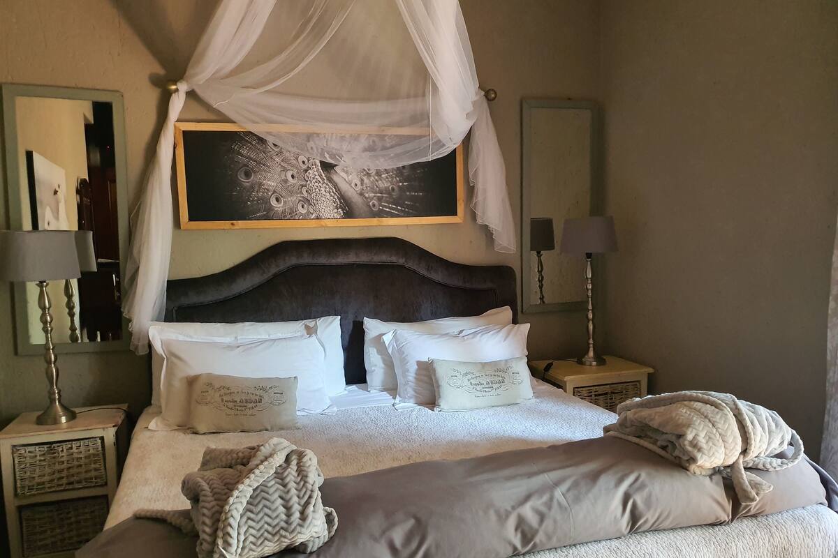 Ein ordentlich gemachtes Bett mit weißen Leinentüchern, einem großen Kopfteil und einem Baldachin. Es gibt zwei Nachttische mit Lampen, einen Spiegel und ein gerahmtes Bild über dem Bett.