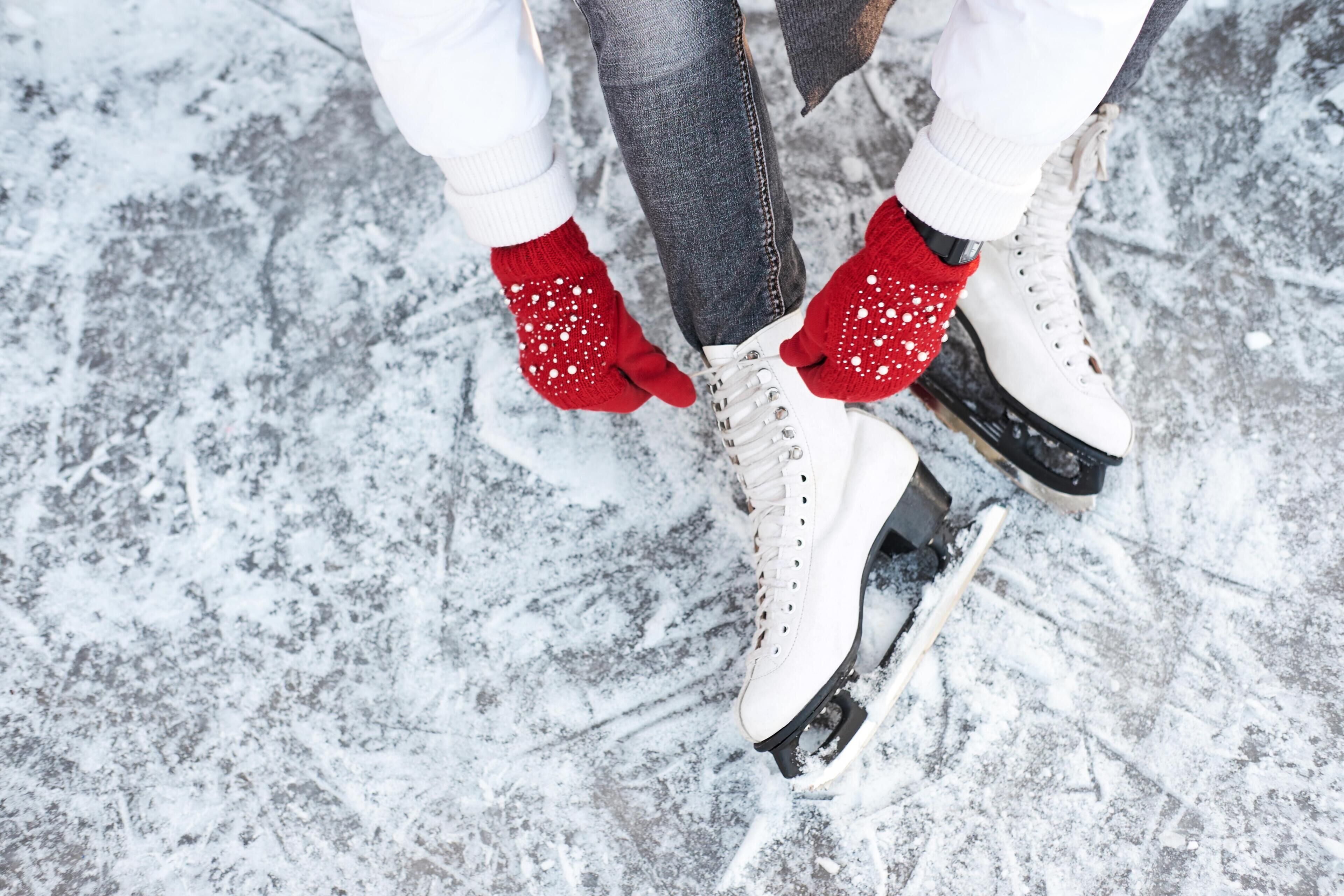 A close-up of a person wearing white ice skates with red gloves on a snowy surface.