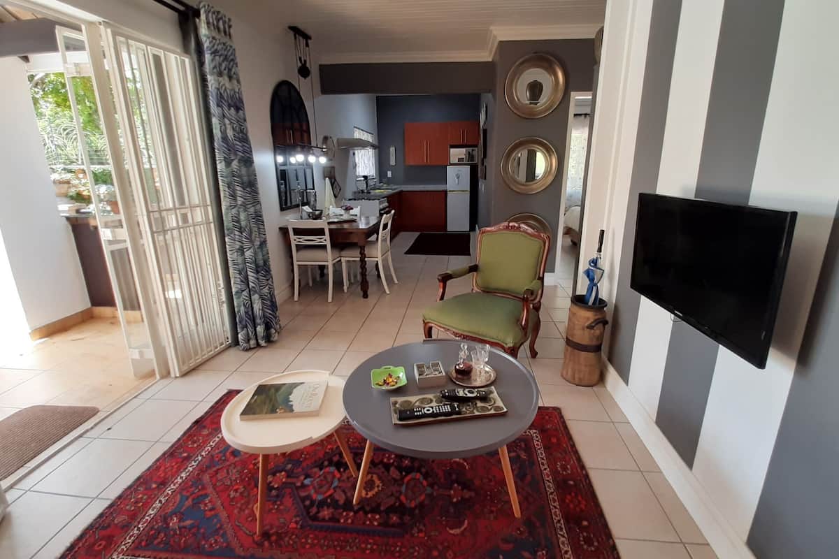 A living room with a TV, a dining area with a table and chairs, a kitchen in the background, and a patterned rug on the floor.