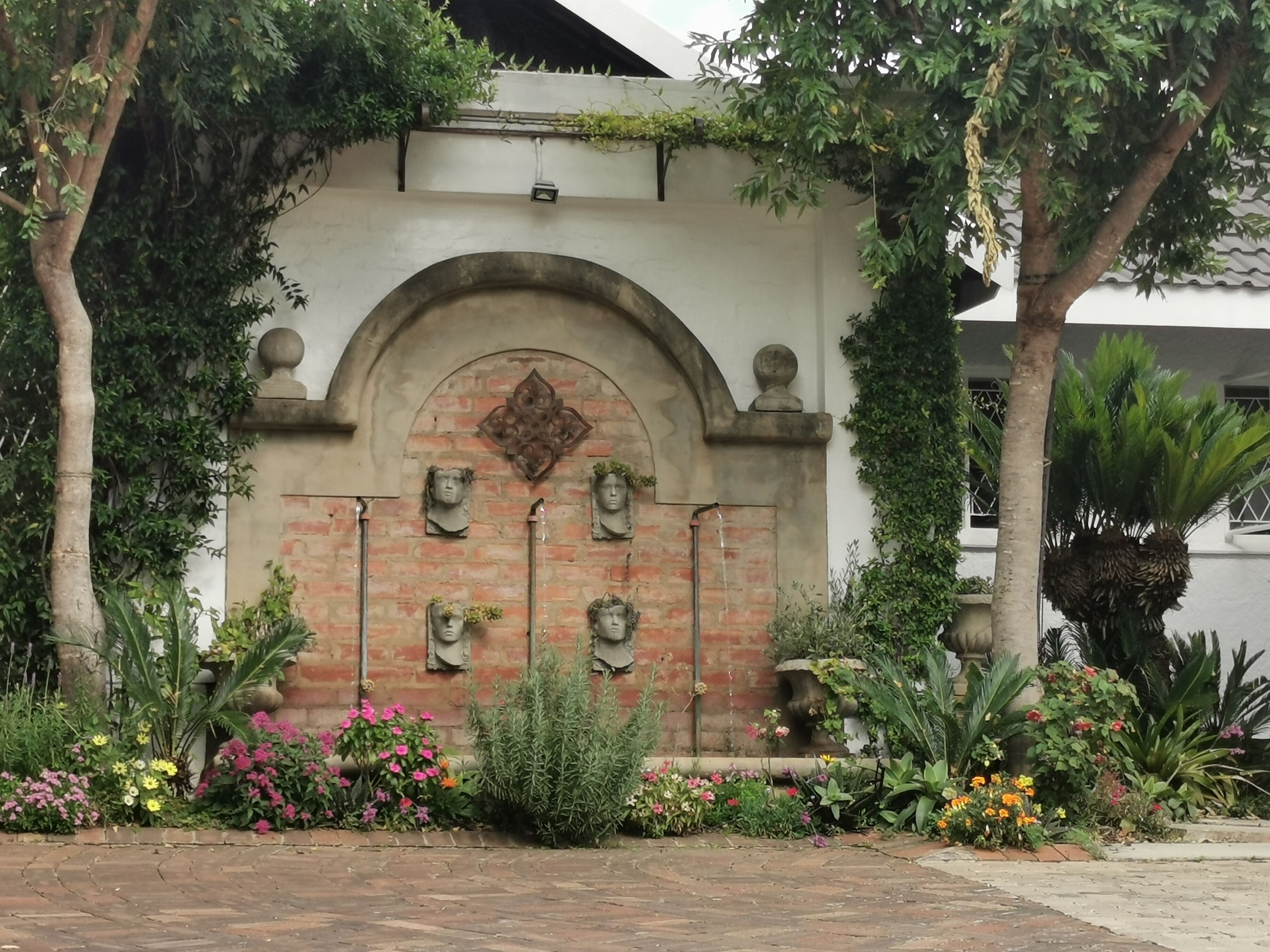 A brick wall with a decorative archway, surrounded by lush greenery and flowering plants.