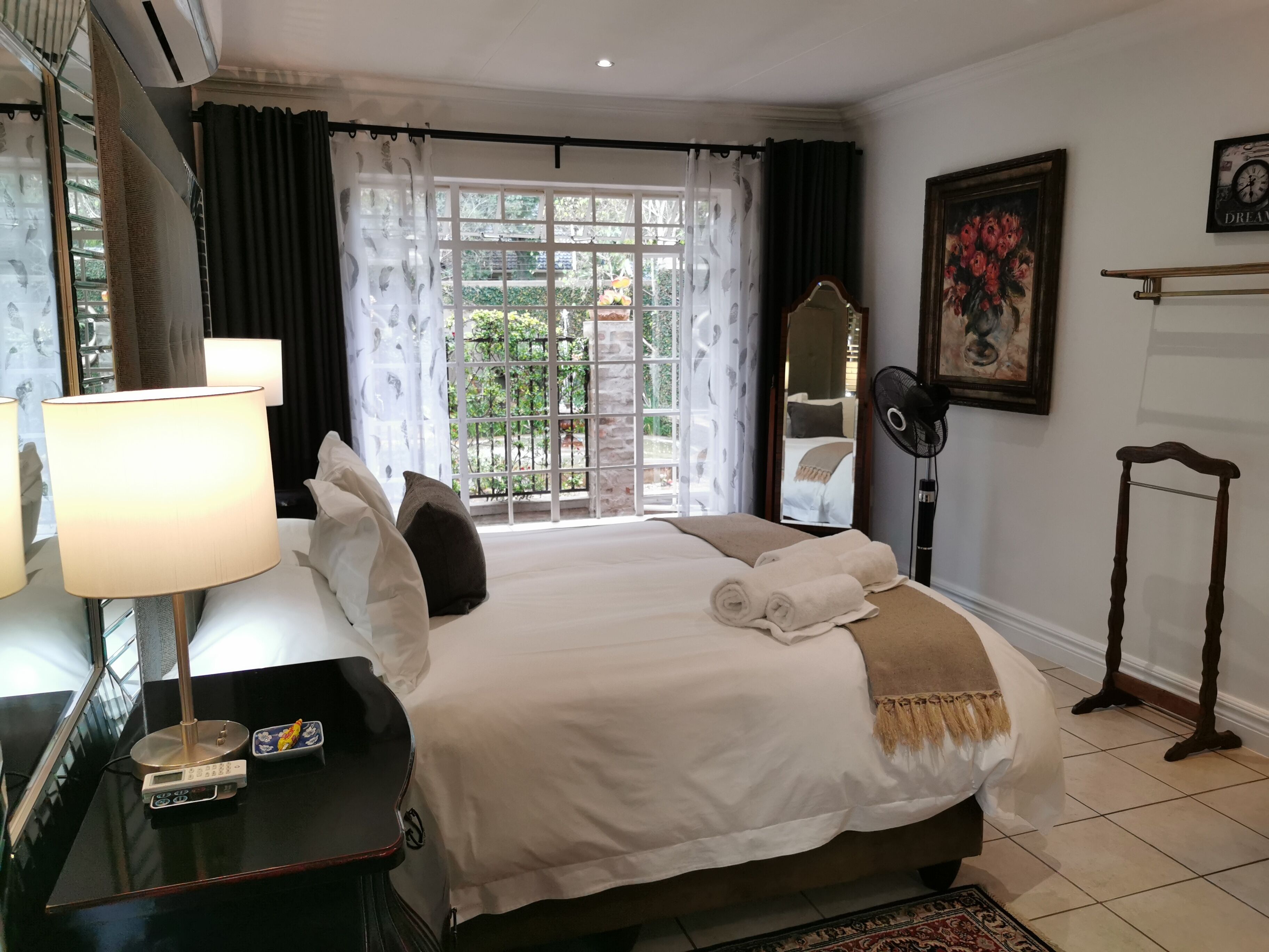 Habitación de hotel con cama, mesita de noche con lámpara, balanza, espejo, ventilador y vista al exterior a través de una ventana.