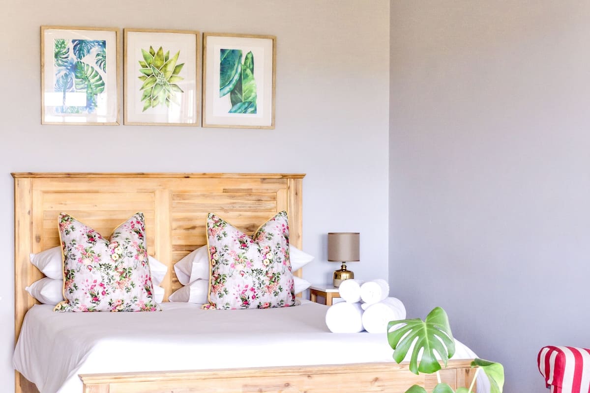 A bedroom with a wooden bed, floral pillows, a bedside lamp, a potted plant, a striped chair, and a rustic chest of drawers.