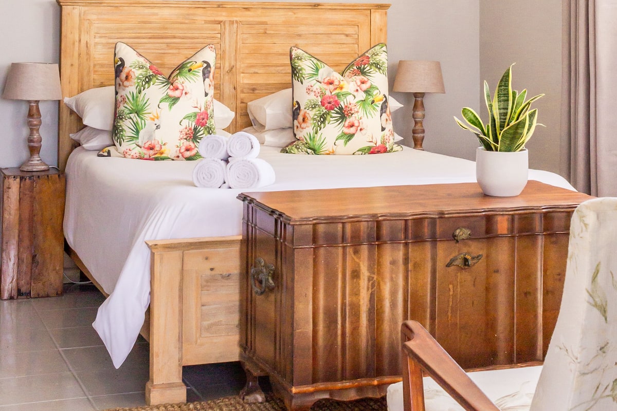 A hotel room with a wooden headboard, two beds with floral pillows, a bedside table with lamps, a small wooden chest, a woven rug, and a potted plant.
