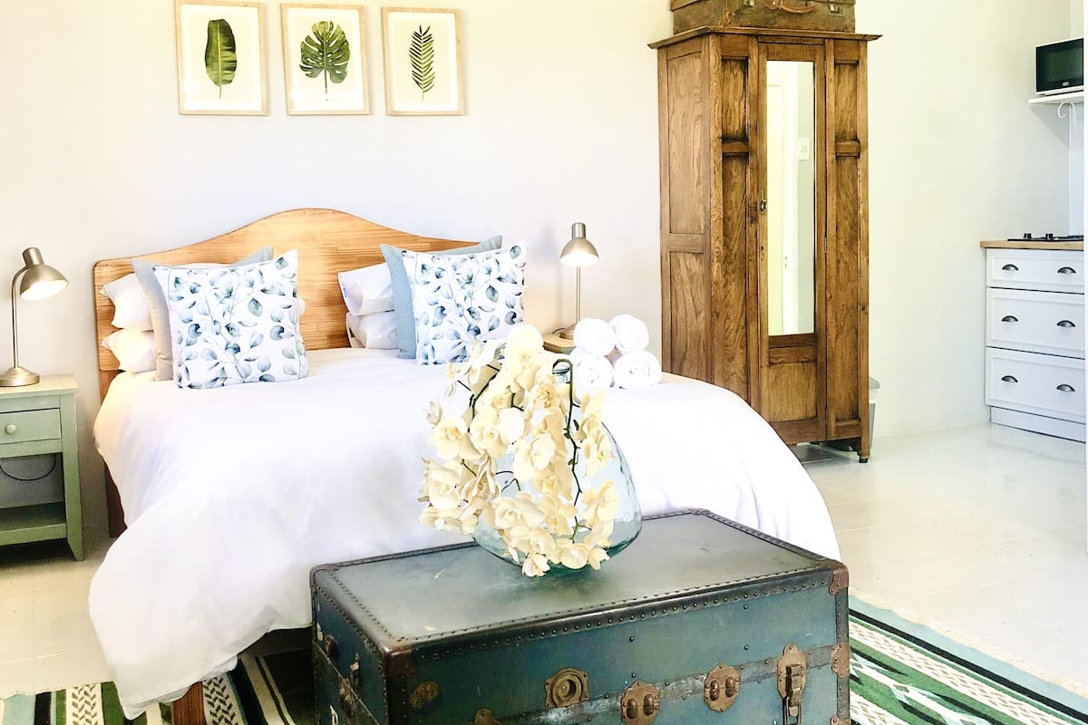 A bedroom with a wooden bed, white bedding, a vintage suitcase with flowers, a wooden wardrobe, and framed leaves on the wall.