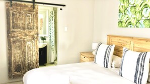 Un dormitorio con cama, cabecera de madera, mesita de noche, baño con lavamanos y espejo, y una puerta con cortina.