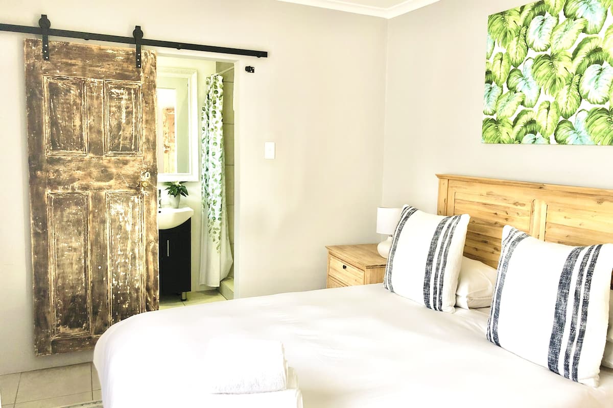 A bedroom with a bed, a wooden headboard, a nightstand, a bathroom with a sink and mirror, and a door with a curtain.