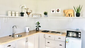 Una cocina moderna con armarios blancos, un lavamanos de acero inoxidable, una tetera blanca y un microondas sobre una encimera blanca. Hay elementos decorativos como un gallo, flores y una planta en maceta en los estantes superiores.