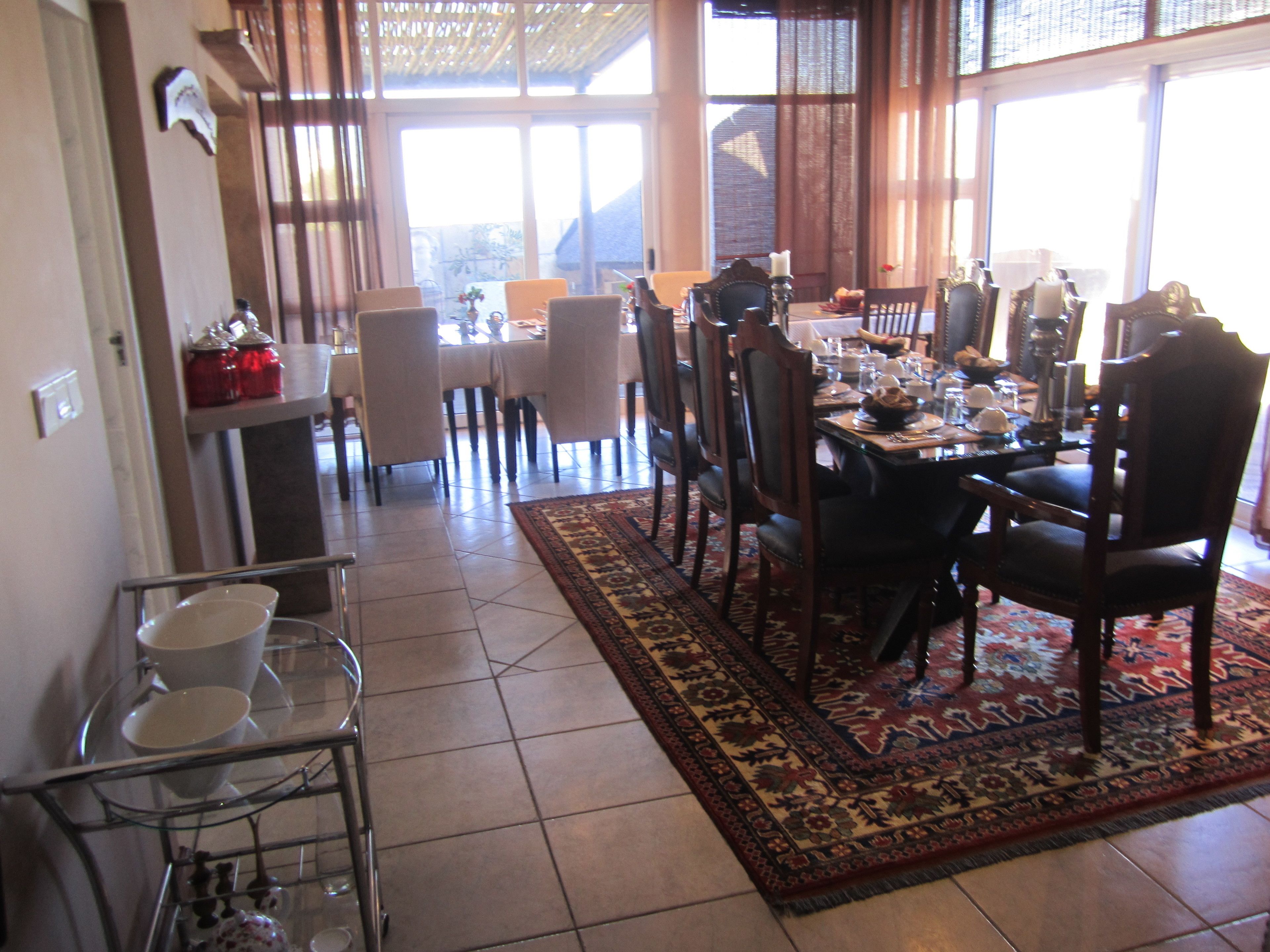 A dining area with a large table, chairs, and a patterned rug. There is a kitchen area with a shelf and jars visible.