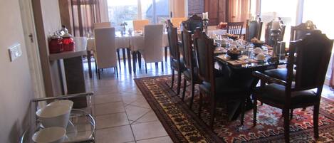 A dining area with a large table, chairs, and a patterned rug. There is a kitchen area with a shelf and jars visible.