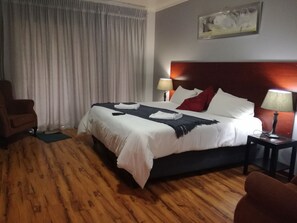 A hotel room with a neatly made bed, a wooden floor, a chair, a nightstand with a lamp, and a painting on the wall. - 21 on Coetzee Guest House (Bloemfontein)
