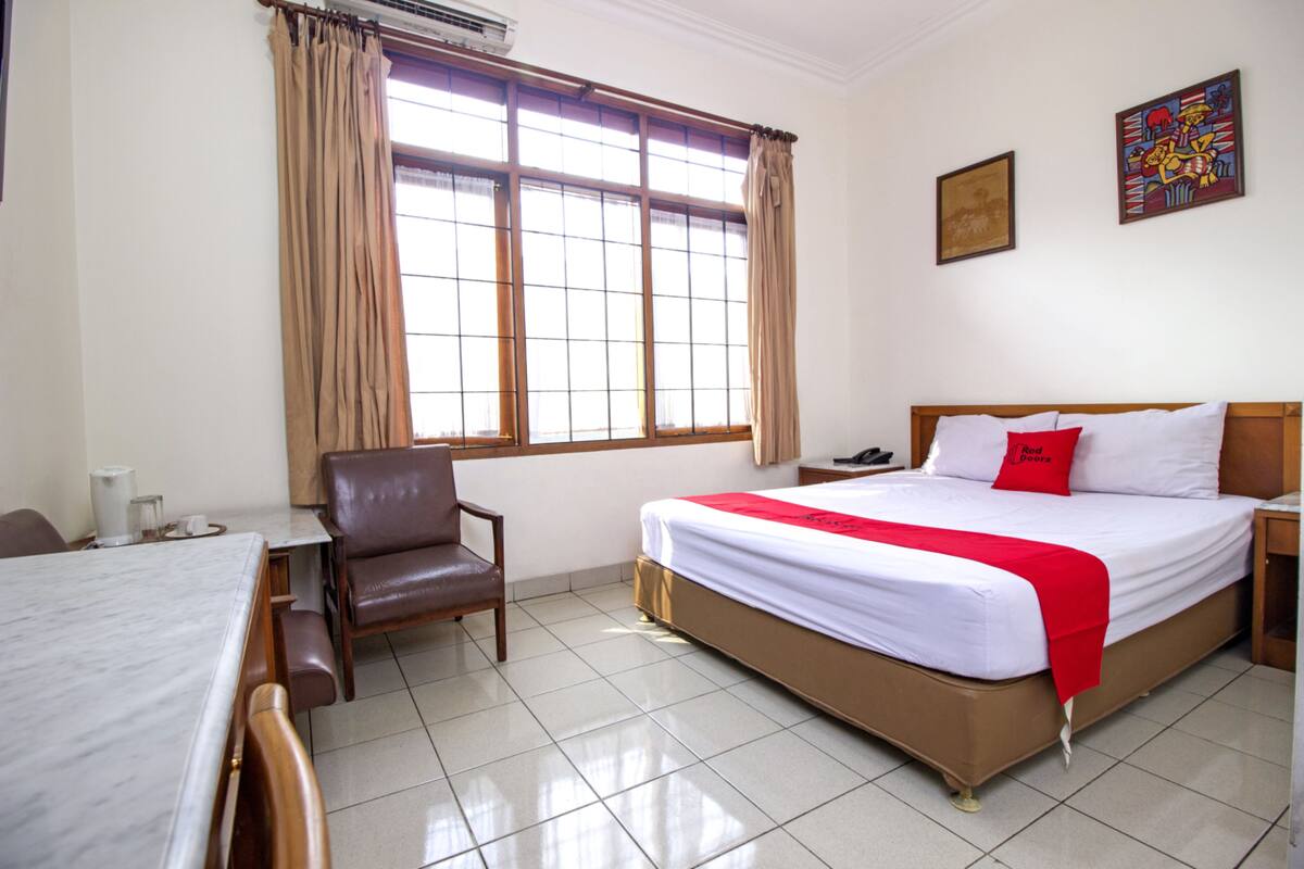 Une chambre d’hôtel comprenant un lit, un bureau, une chaise, une fenêtre avec des rideaux et un mur orné de deux tableaux.
