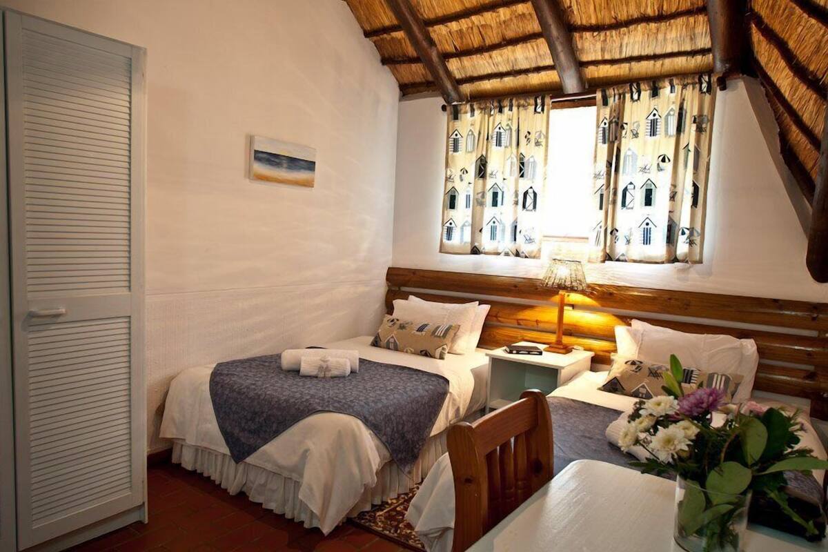 A cozy bedroom with two beds, a wooden headboard, a window with patterned curtains, and a bedside table with a lamp.
