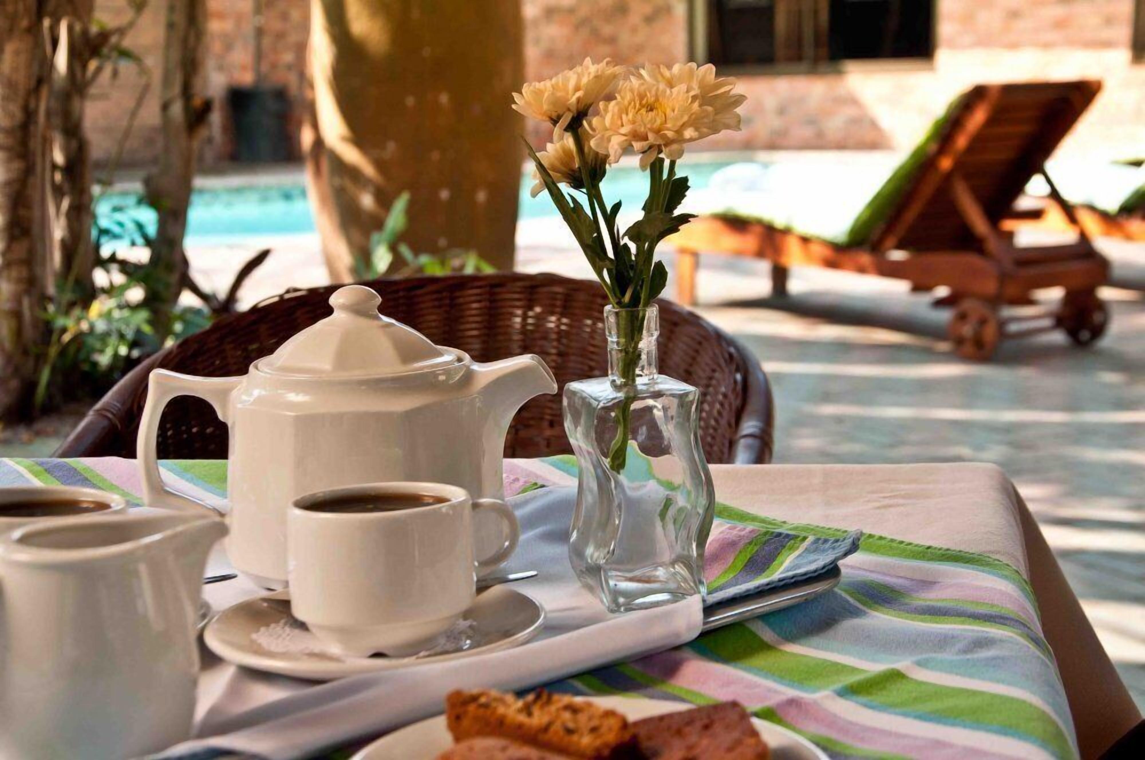 A table with a tray containing a teapot, cups, and a vase with flowers, set outdoors with a pool and lounge chairs in the background.