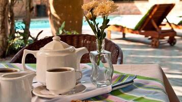 A table with a tray containing a teapot, cups, and a vase with flowers, set outdoors with a pool and lounge chairs in the background.