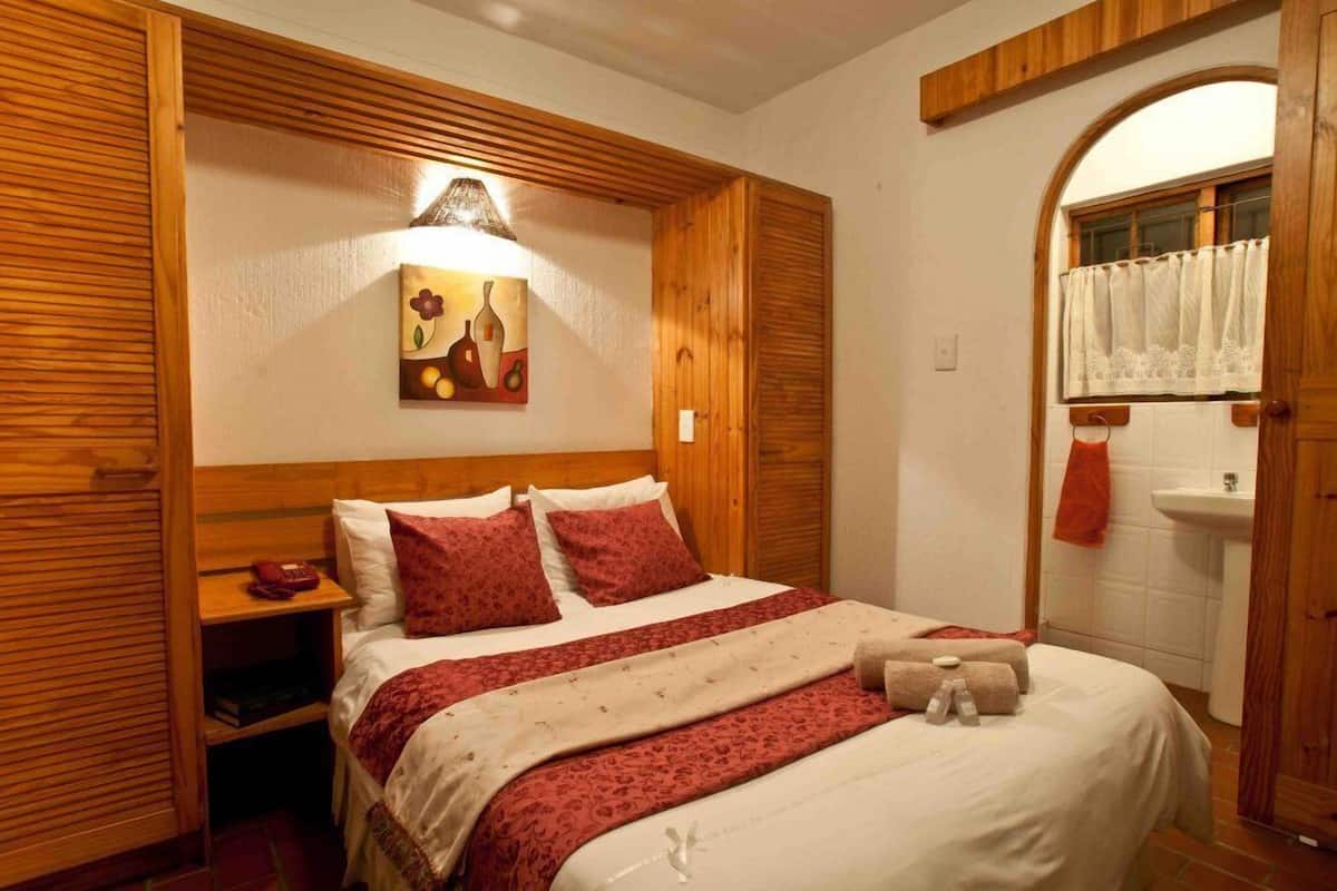 A hotel room with a wooden headboard, a bed with red and white bedding, a painting on the wall, and a bathroom with a sink and a window with a curtain.