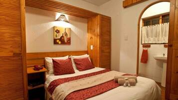 A hotel room with a wooden headboard, a bed with red and white bedding, a painting on the wall, and a bathroom with a sink and a window with a curtain.